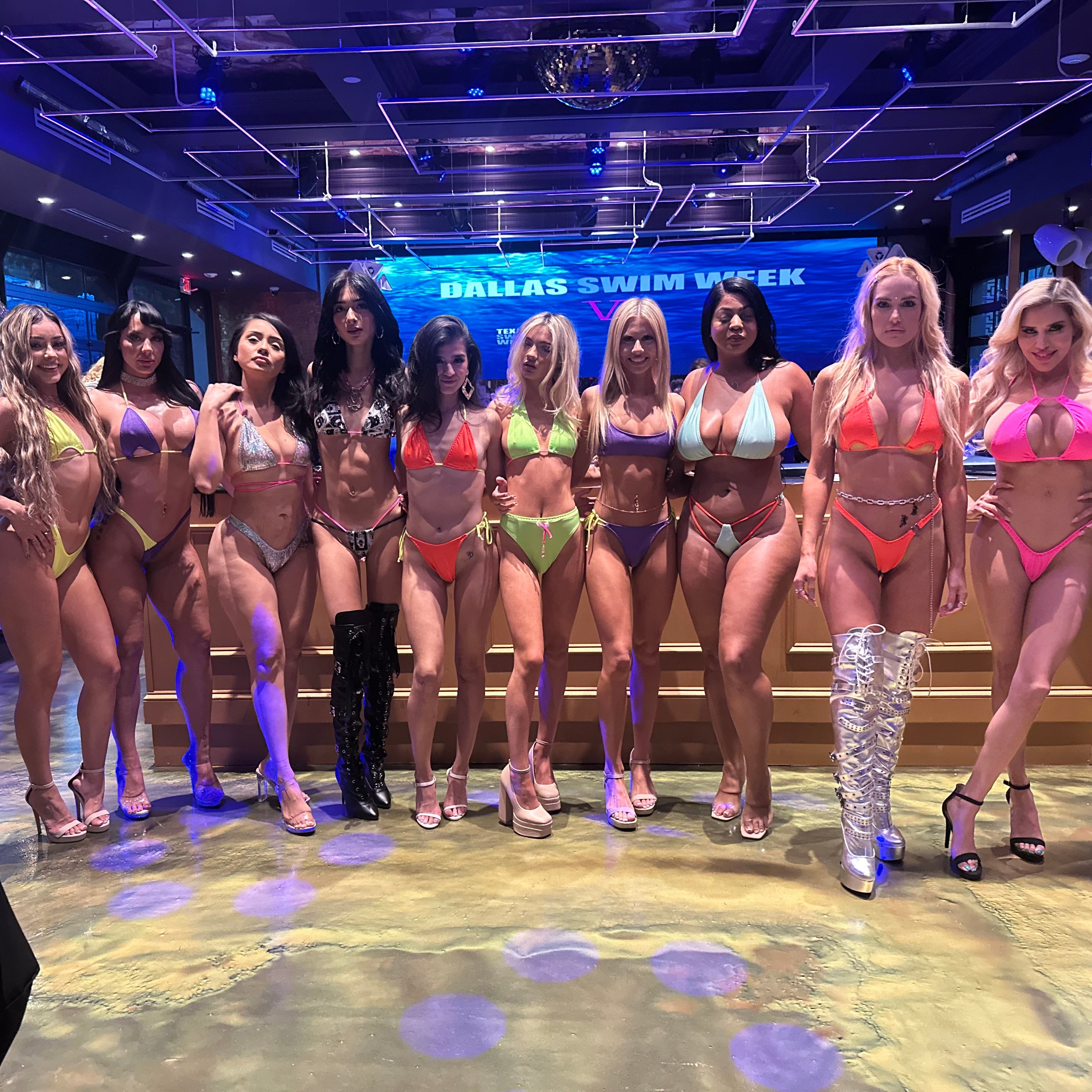 A group of ten women posing together in various colorful bikinis and high-heeled sandals on a stage-like floor with a 'Dallas Swim Week' sign in the background.