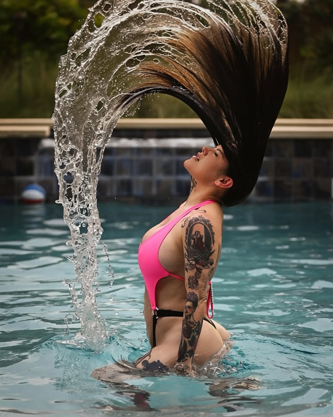 A woman in a pink and black bikini stands in a swimming pool with her hair dramatically tossed into the air, creating a large splash. She has visible tattoos on her arm and shoulder.